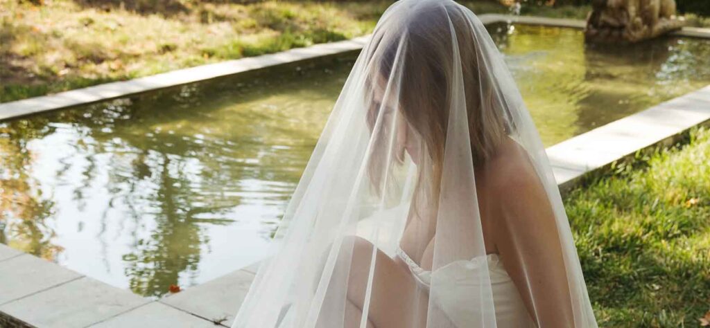 Bride sitting by a reflective pool in a white wedding dress and veil, outdoors in a garden setting.