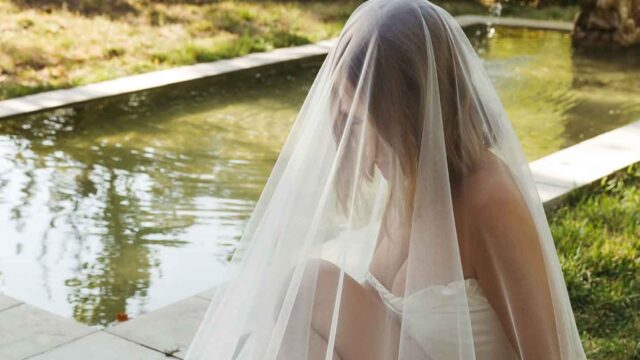 Bride sitting by a reflective pool in a white wedding dress and veil, outdoors in a garden setting.