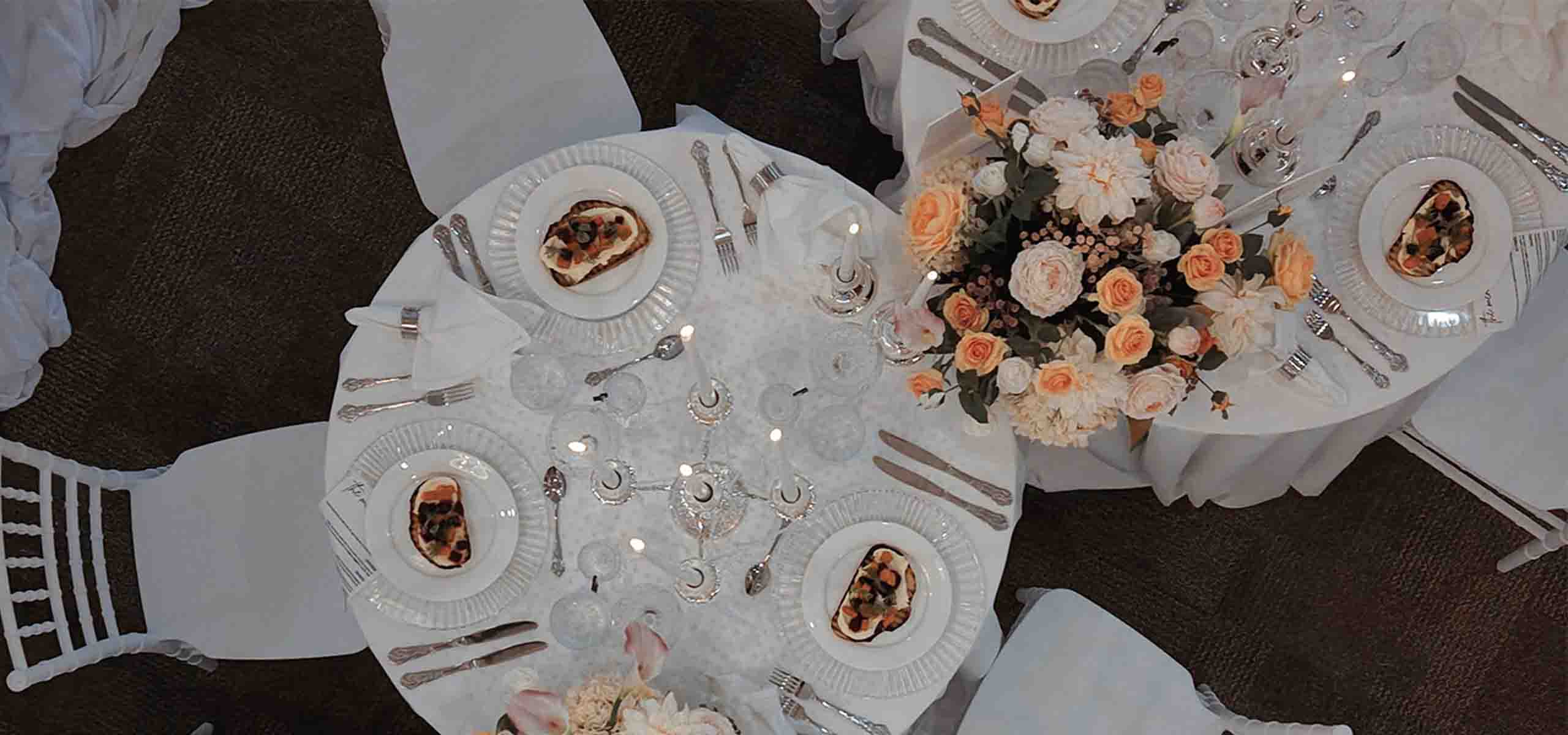Overhead view of a formal round table set for dinner with white china, silverware, candles, and a peach-and-cream floral centerpiece.