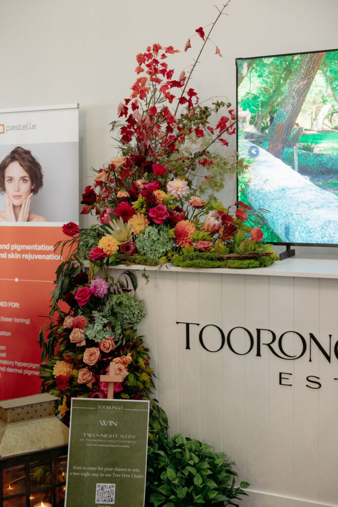 Decorative flower arrangement with red, pink, and peach blooms beside a digital display screen in a showroom.