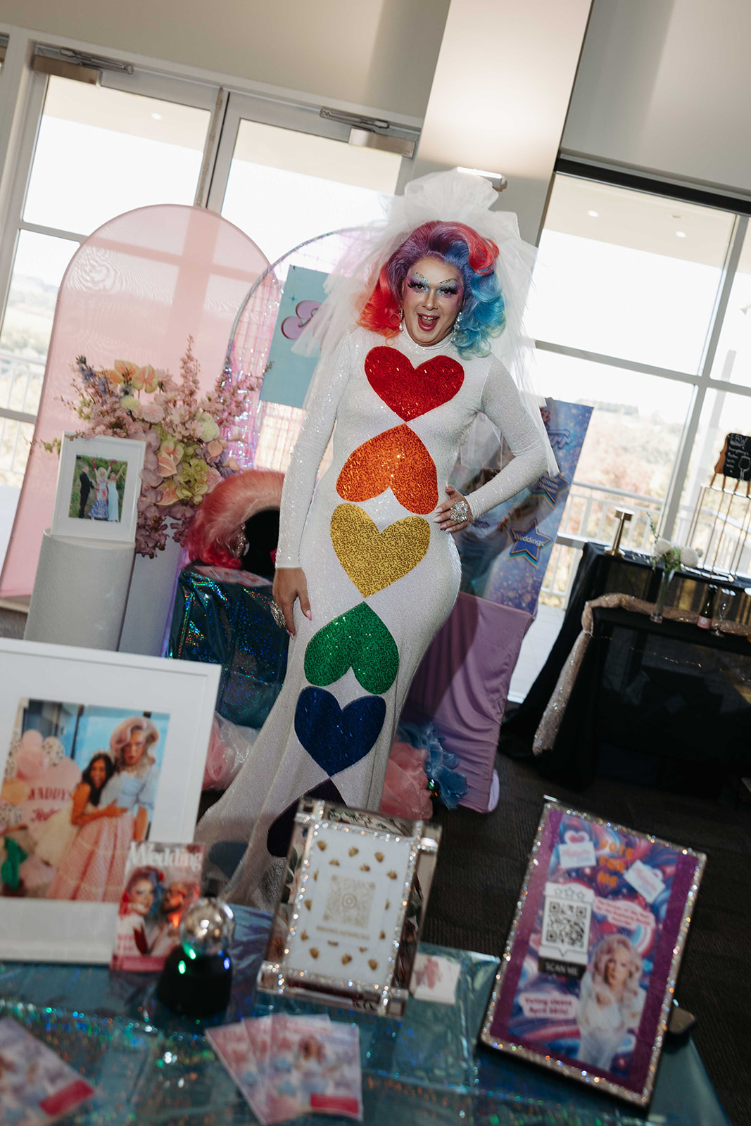 Person in a glittery white gown with rainbow heart shapes down the front, wearing a bright rainbow wig and veil at a party setup.