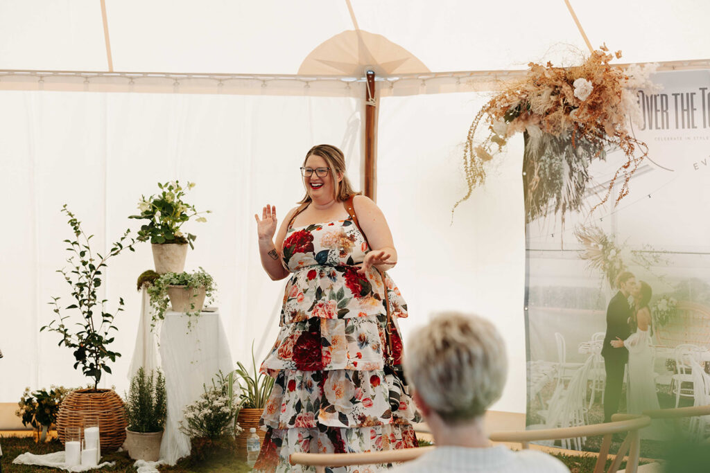 Plus-size woman in a floral dress speaking on a decorated stage with potted plants and a wedding backdrop nearby.