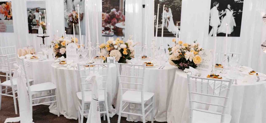 Elegant white-themed wedding reception table with candles and pastel floral centerpieces on a long, linen-draped table.