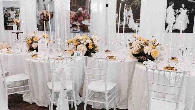 Elegant white-themed wedding reception table with candles and pastel floral centerpieces on a long, linen-draped table.