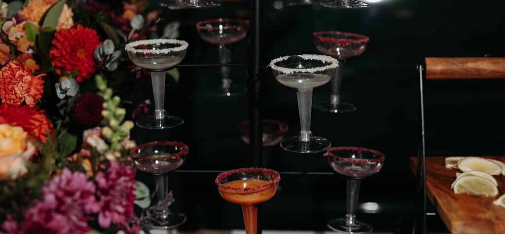 Coupe glasses with sugared rims arranged on a glass rack, an orange cocktail in the foreground, and lemon wedges on a wooden board to the right, set against dark background with colorful flowers on the left.