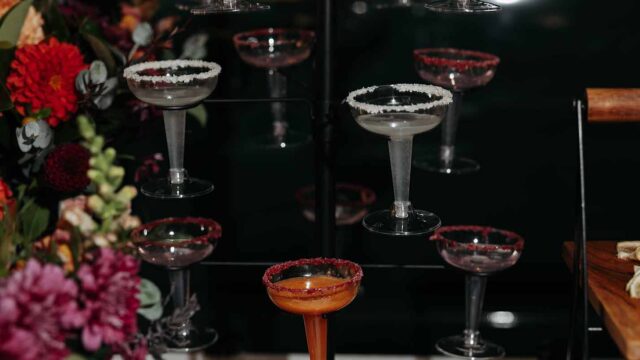 Coupe glasses with sugared rims arranged on a glass rack, an orange cocktail in the foreground, and lemon wedges on a wooden board to the right, set against dark background with colorful flowers on the left.