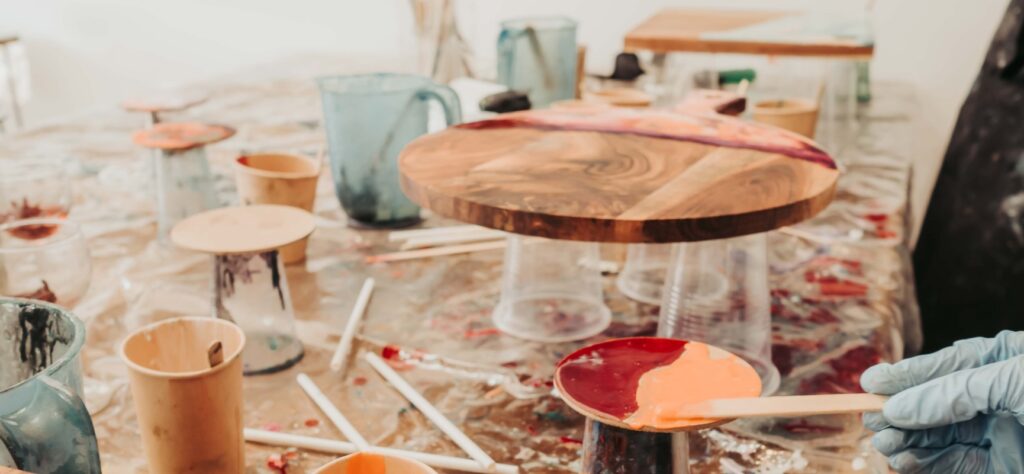 Gloved hand holding a wooden palette with orange paint in a cluttered painting studio with cups and brushes around it