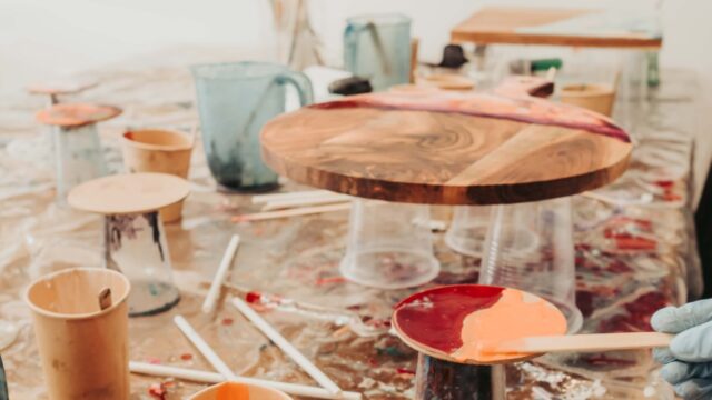 Gloved hand holding a wooden palette with orange paint in a cluttered painting studio with cups and brushes around it
