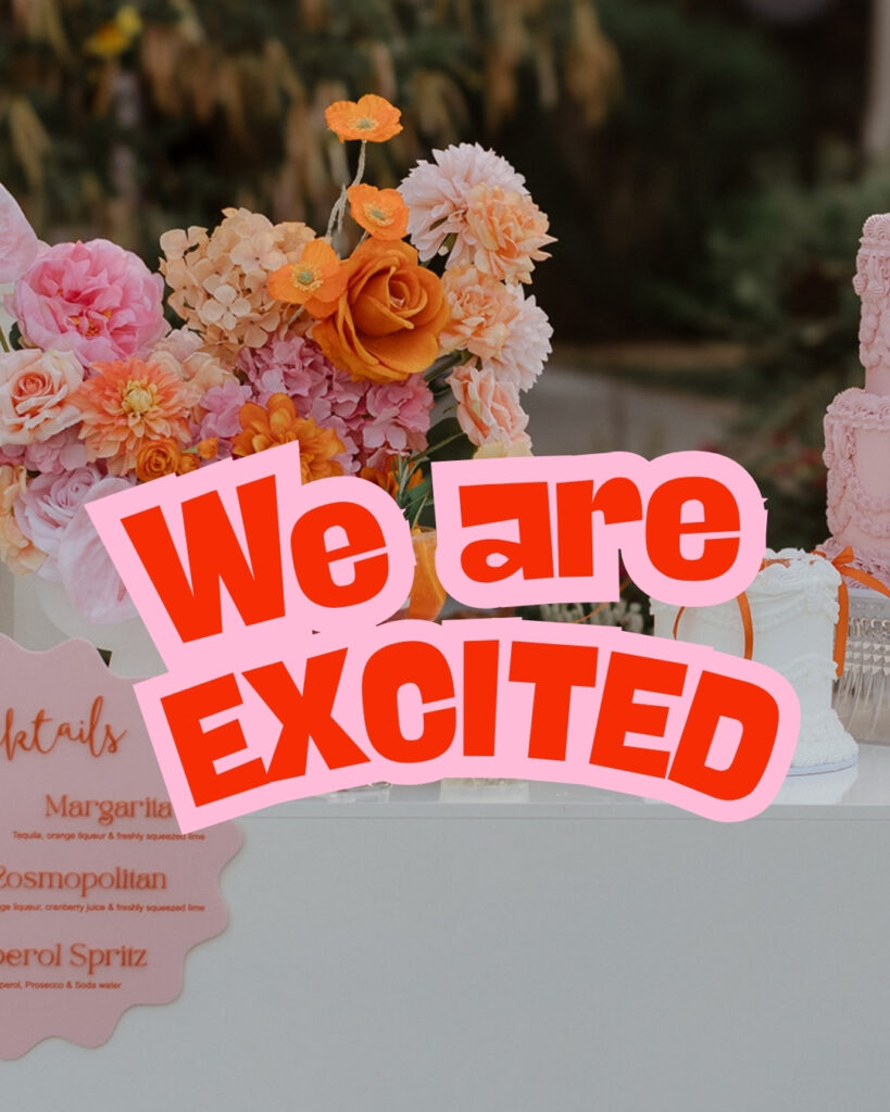 Decorative dessert table with peach/pink flowers and a pink banner reading 'We are EXCITED' across the foreground.