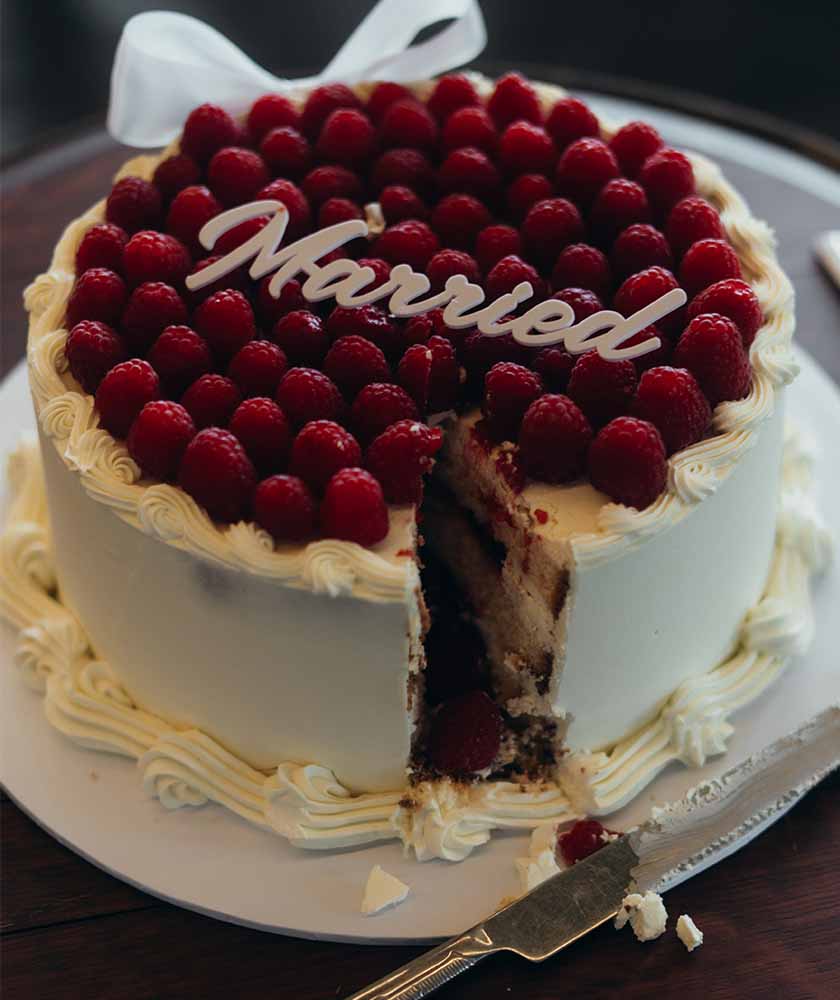 Round cake with white icing and piped borders, topped with fresh raspberries; a slice has been cut out.