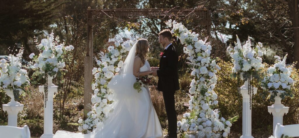 Bride in a white gown and groom in a dark suit hold hands under a white floral arch outdoors during their ceremony.