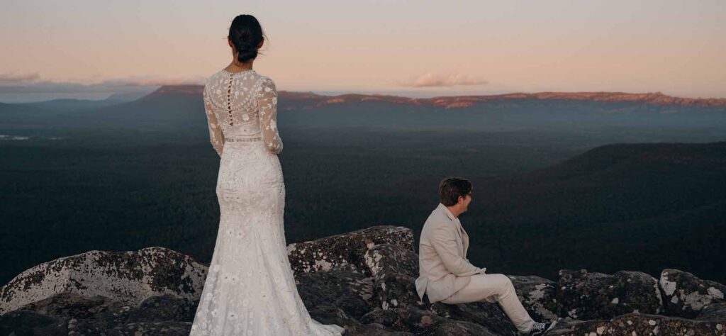 Bride in a lace wedding dress stands on a rocky cliff, facing a sunset with a man in a light suit seated nearby on the rocks.