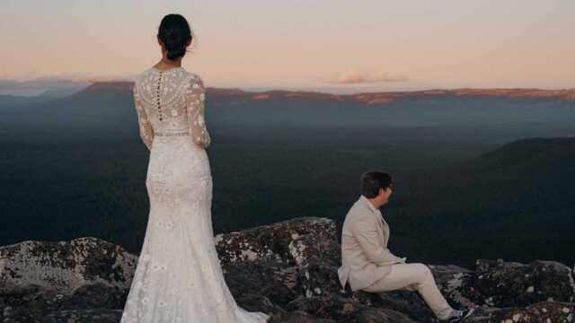 Bride in a lace wedding dress stands on a rocky cliff, facing a sunset with a man in a light suit seated nearby on the rocks.