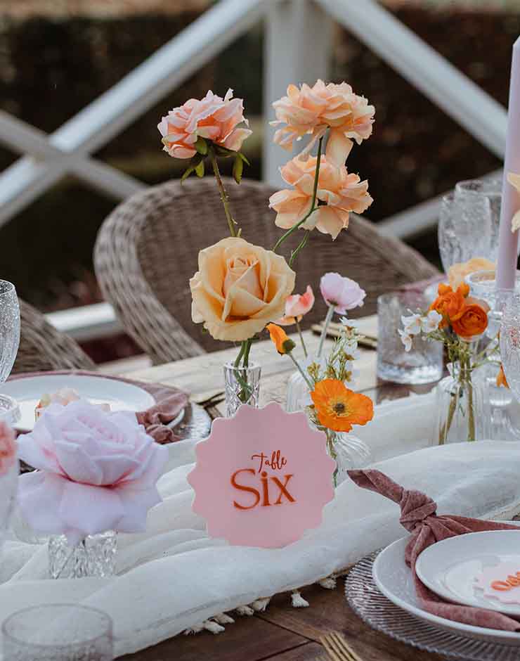 1 - I do I do Gippsland Table centerpiece with peach roses in glass vases and a pink 'Table Six' card on a white table runner.