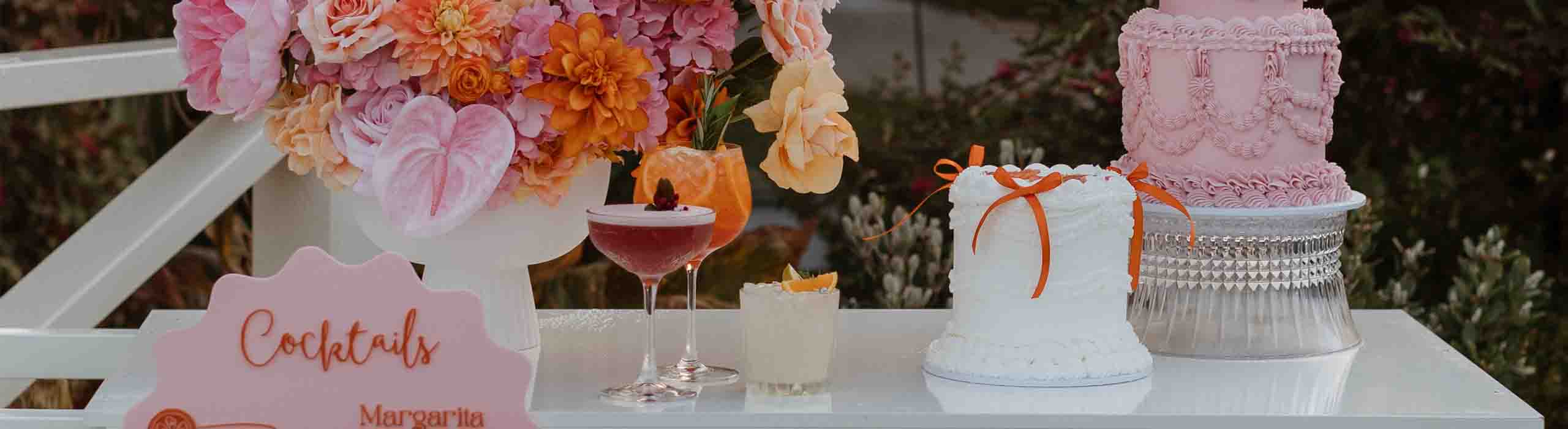 1 - I do I do Gippsland Table dessert display with a bright pink floral arrangement, two cocktails, a white cake with orange ribbon, and a pink frosted tiered cake.
