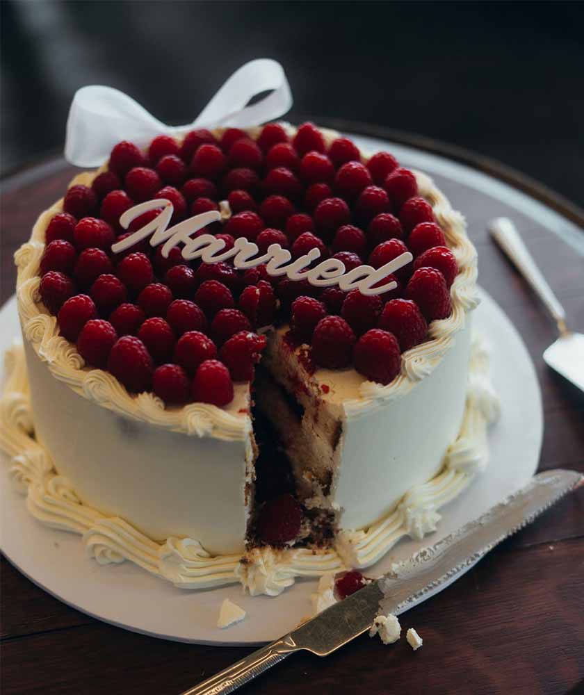 Wedding cake topped with raspberries and a white chocolate 'Married' script, a slice removed for serving.