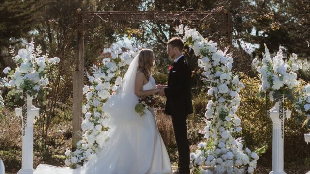 Bride in a white gown and groom in a dark suit hold hands under a white floral arch outdoors during their ceremony.