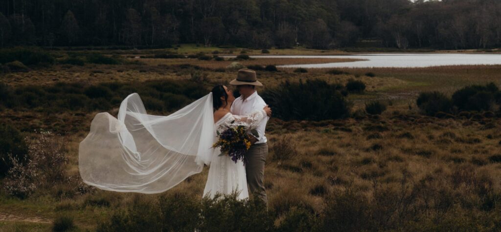 Bride in lace gown and groom in a hat embrace in a field, veil billowing between them with a bouquet in the foreground