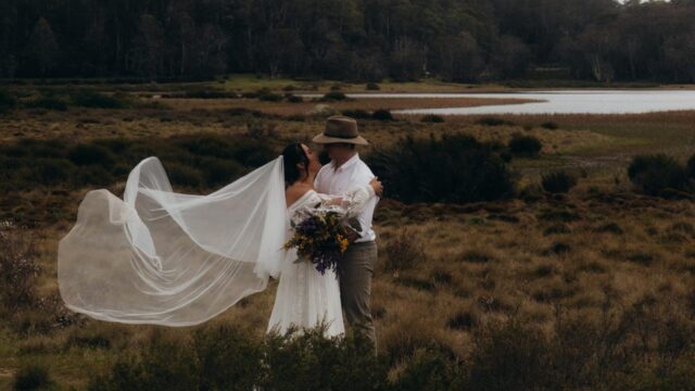 Bride in lace gown and groom in a hat embrace in a field, veil billowing between them with a bouquet in the foreground