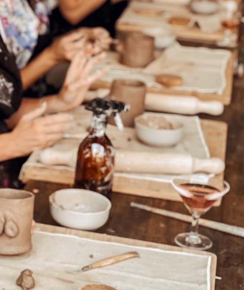 Hands shaping clay at a pottery workshop, with bowls, rolling pins, and a spray bottle on the table.