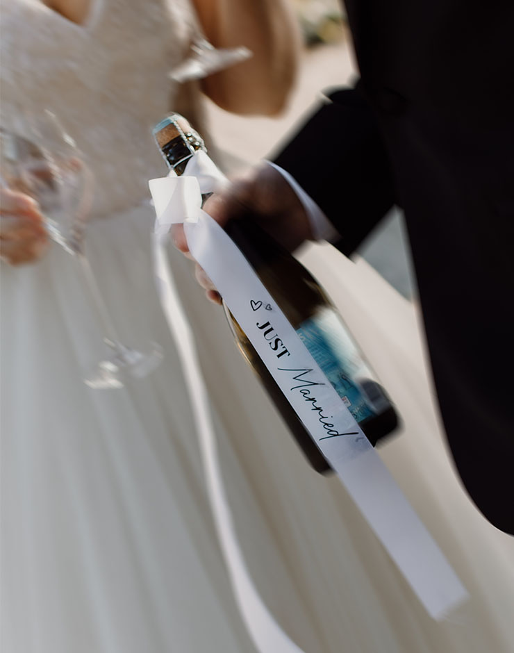 2 - I do I do Gippsland Bride and groom holding a champagne bottle with a white'Just Married' ribbon in focus, sugaring the celebration moment.