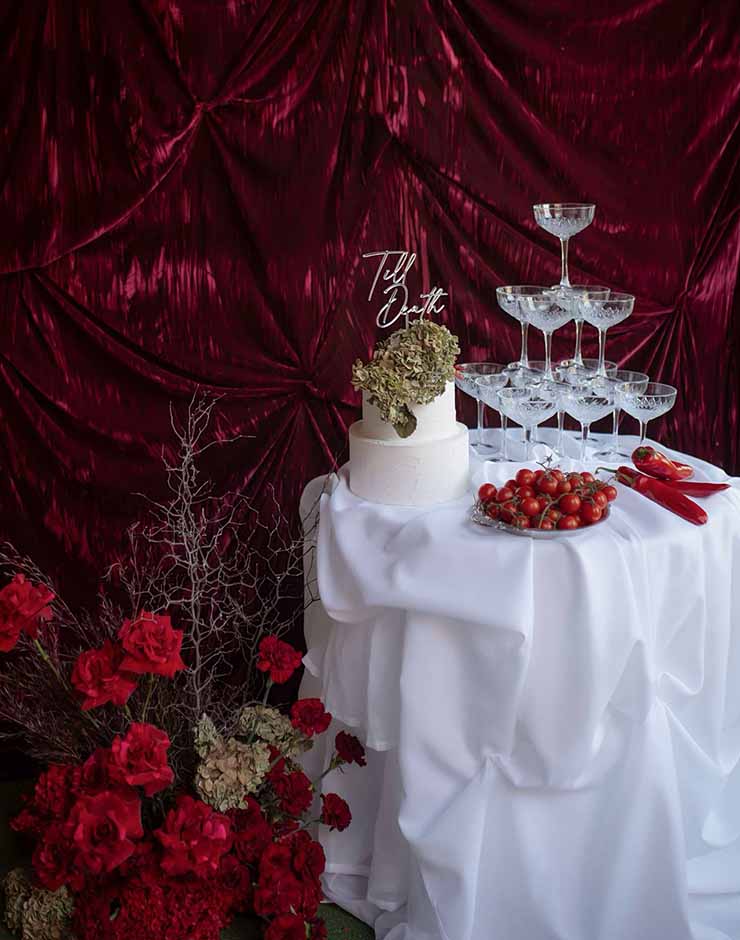 2 - I do I do Gippsland White wedding cake with a dried floral topper on a white-draped table beside a champagne-glass tower and a bowl of cherry tomatoes against a burgundy backdrop.
