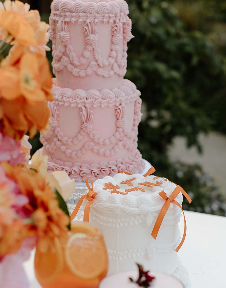 2 - I do I do Gippsland Three-tier pink buttercream cake with elaborate piping, on a dessert table with a white cake accented by orange ribbons and orange flowers nearby.