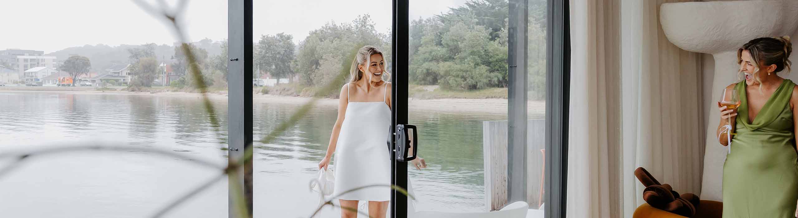 Two women at a lakeside venue: one in a white dress steps through sliding glass doors as the other, in a green dress, drinks wine indoors and laughs.