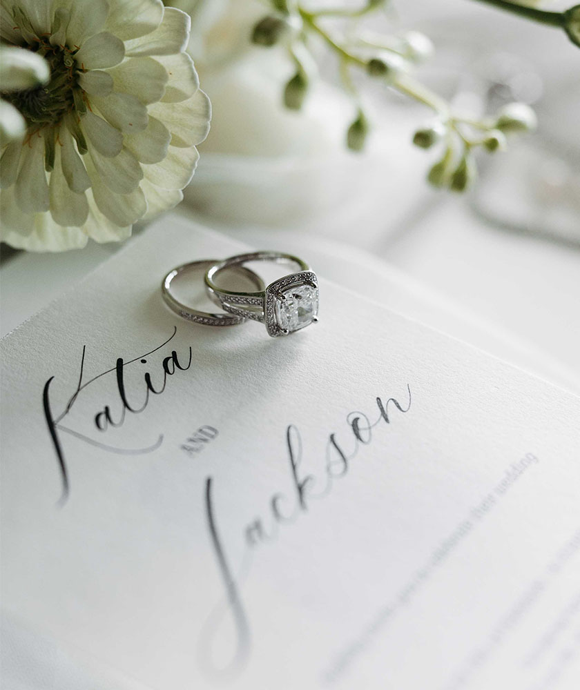 Two silver rings (engagement and wedding band) rest on a white wedding invitation with elegant script and a pale flower nearby, suggesting a wedding scene.