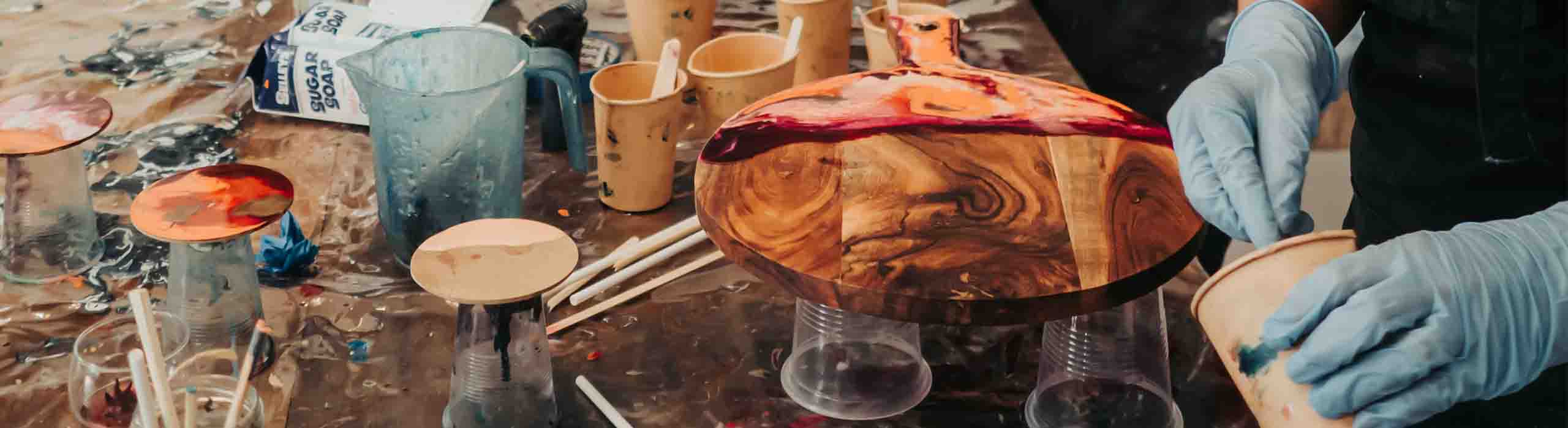 Artist wearing blue gloves works on a polished wood bowl with colorful resin on a messy studio table