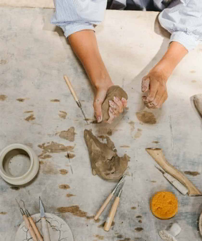 Hands kneading wet clay on a pottery worktable with chisels and carving tools nearby.