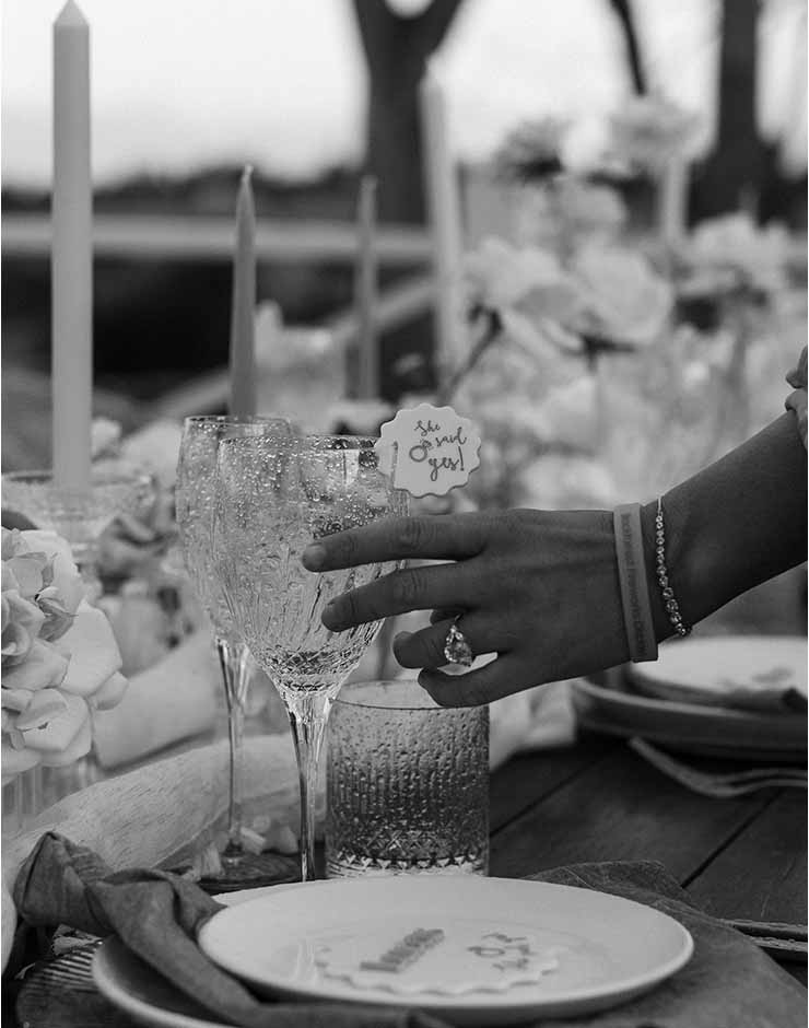 3 - I do I do Gippsland A guest's hand with bracelets and a ring reaches for a crystal wine glass at an outdoor wedding table with candles and flowers (black and white).