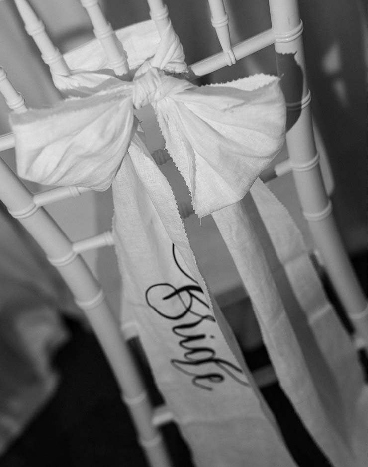 3 - I do I do Gippsland White chair decorated with a tied fabric bow and a hanging ribbon that reads 'Bride' for a wedding.