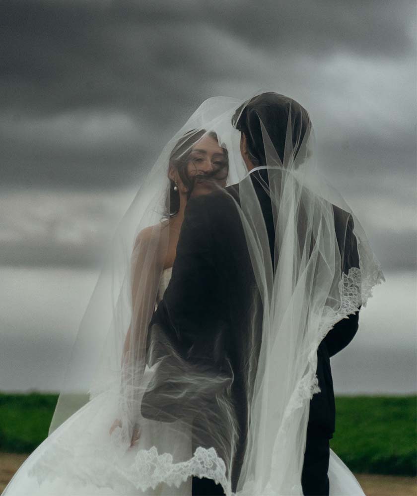 Bride and groom stand close together, their faces near under a long veil on a windy, overcast day with a green field in the background.