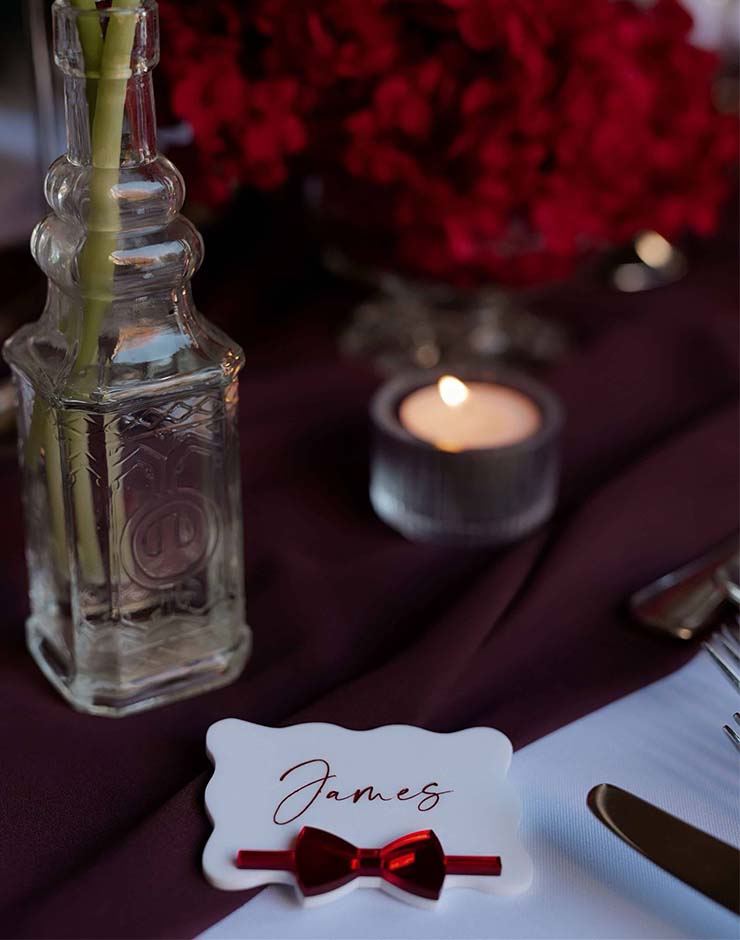 3 - I do I do Gippsland Place card reading 'James' with a red bow on a burgundy tablecloth beside a lit candle and glass centerpiece.