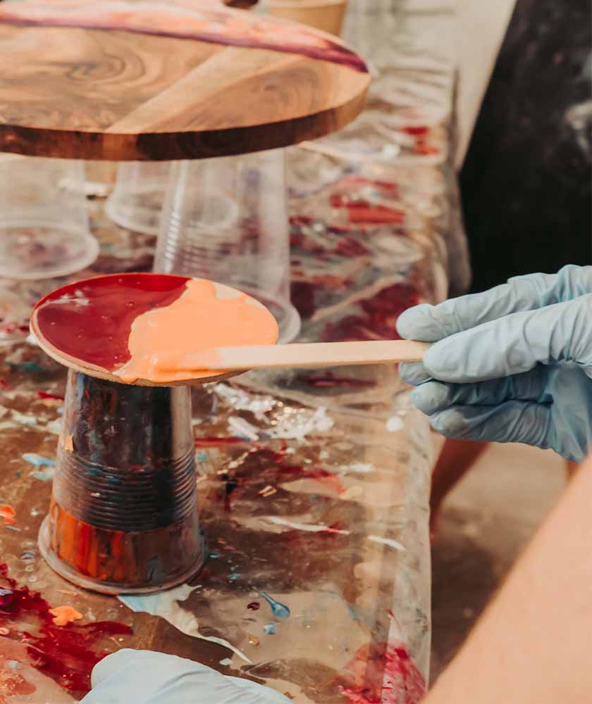 Gloved hand uses a wooden stick to spread orange paint onto a wooden spoon over a paint-splattered workspace.
