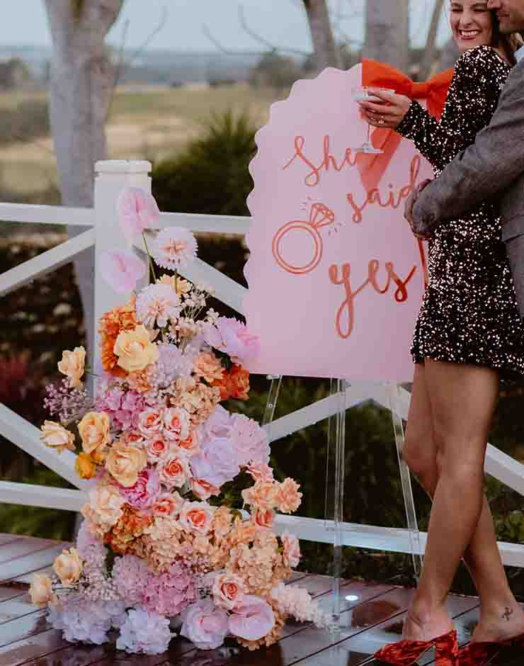 4 - I do I do Gippsland Couple toasting with champagne beside a pink 'she said yes' sign and a large pastel flower arrangement on a wooden deck.