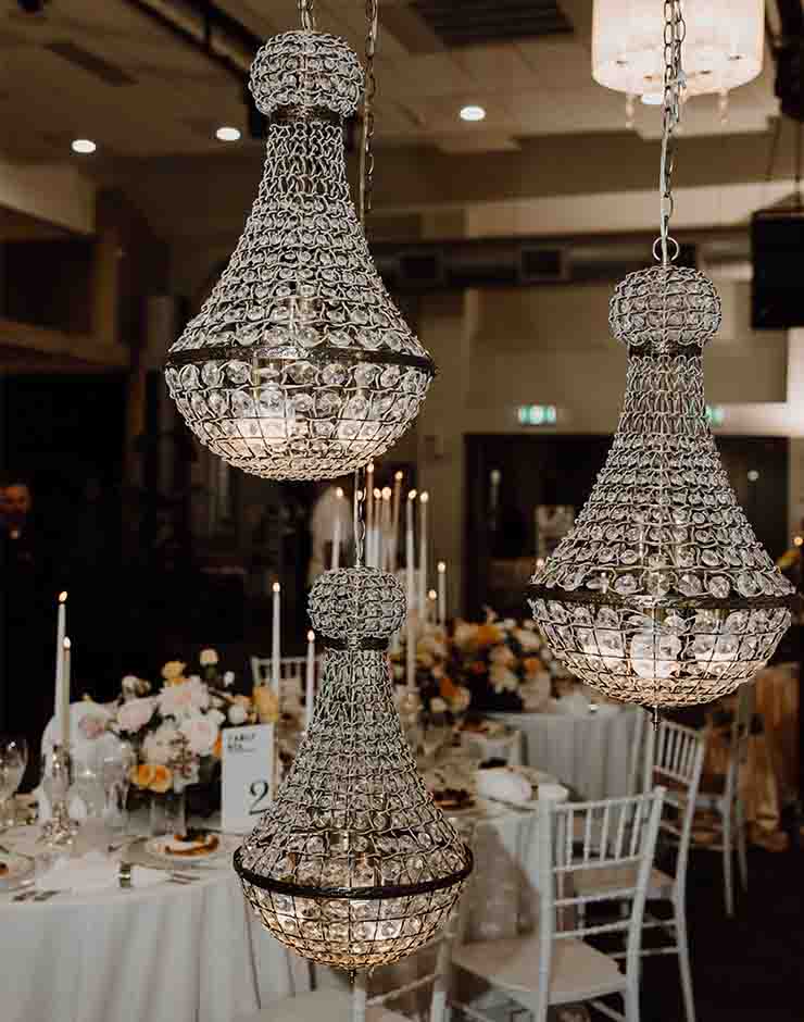 5 - I do I do Gippsland Crystal chandeliers hanging over a wedding reception with round tables set with white linens and flowers on the tables.