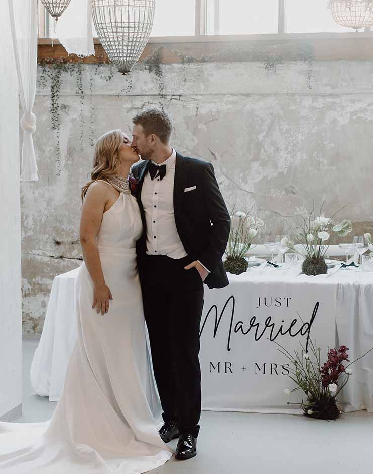 5 - I do I do Gippsland Bride and groom in wedding attire kiss beside a cake table with a 'Just Married' banner