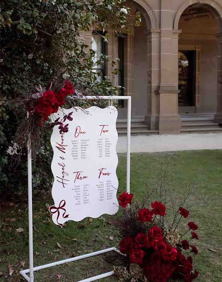 5 - I do I do Gippsland Outdoor wedding seating chart on a white frame, surrounded by red roses, in front of a stone building.