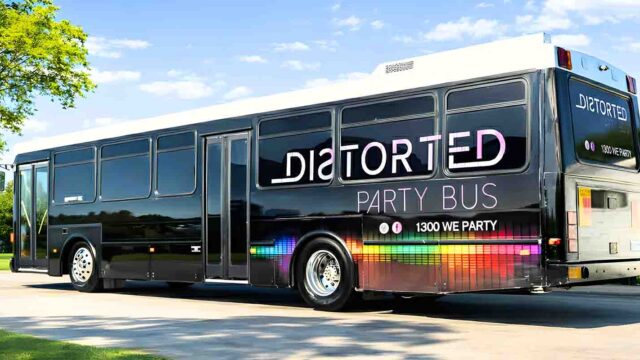 Black party bus with rainbow light-strip graphics and large white lettering, parked on a sunny driveway beside trees and a lake.