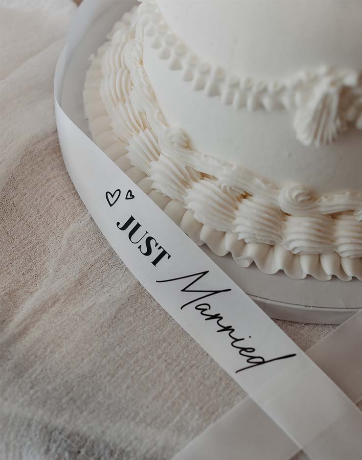 6 - I do I do Gippsland White frosted wedding cake with decorative piped frosting and a 'Just Married' ribbon draped around it.