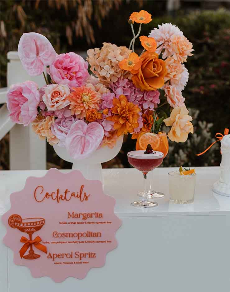 7 - I do I do Gippsland Elegant cocktail setup with a large peach-pink floral arrangement in a white vase on a bar, plus two glasses and a pink cocktail sign listing margarita, cosmopolitan, and Aperol Spritz