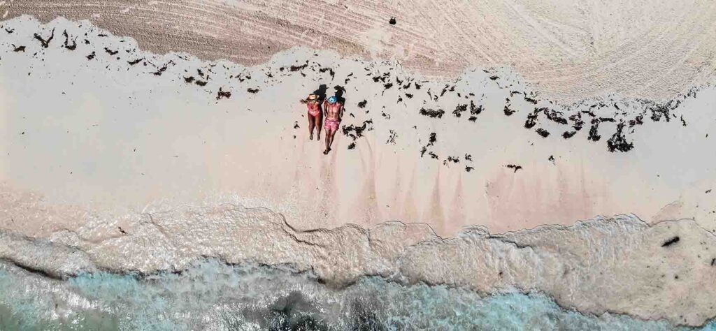 Two people in pink swimsuits sunbathing on a sandy beach, viewed from above with turquoise water nearby.