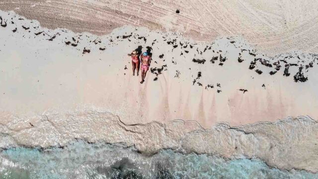 Two people in pink swimsuits sunbathing on a sandy beach, viewed from above with turquoise water nearby.