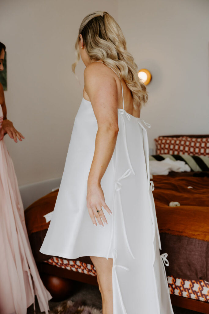 Woman in a white dress standing with back to camera in a bedroom, showing tied back details of her gown.
