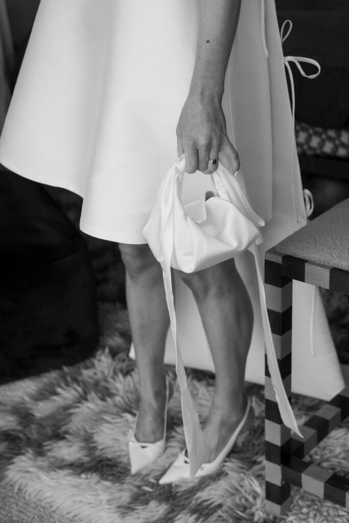 Woman's hand grips a white gathered clutch, matching a white dress and pointed heels, standing on a shaggy rug beside a striped table