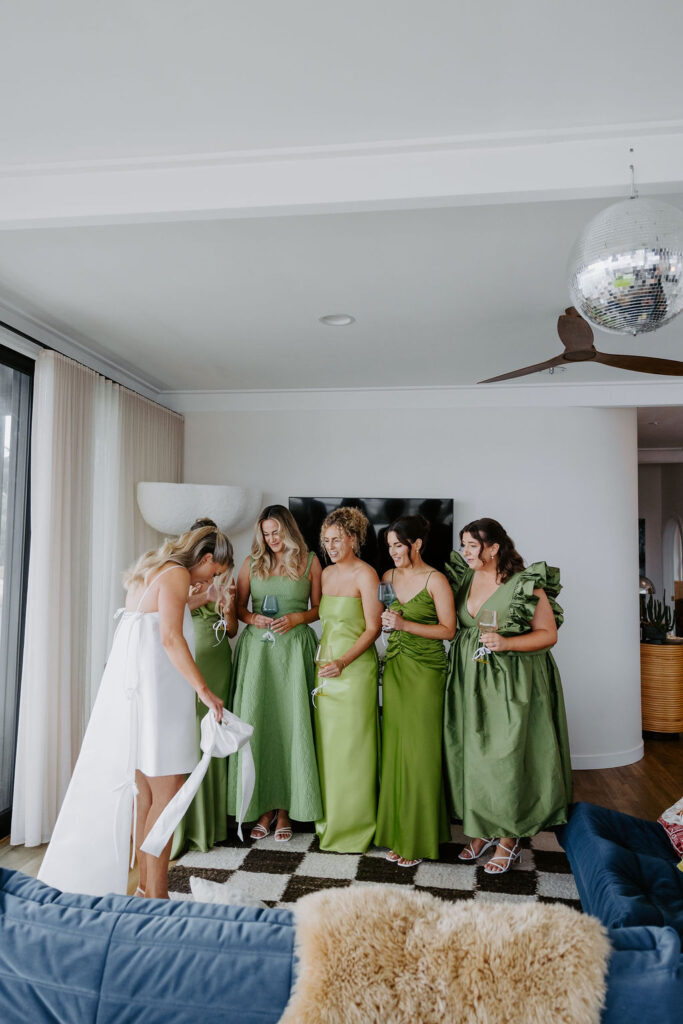 Bride in a white wedding dress stands with six bridesmaids in green gowns, holding wine glasses in a bright living room with a checkered rug and sofa.