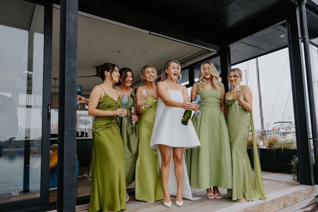 Bride in a white dress toasts with champagne as six bridesmaids in green gowns cheer on a yacht deck.