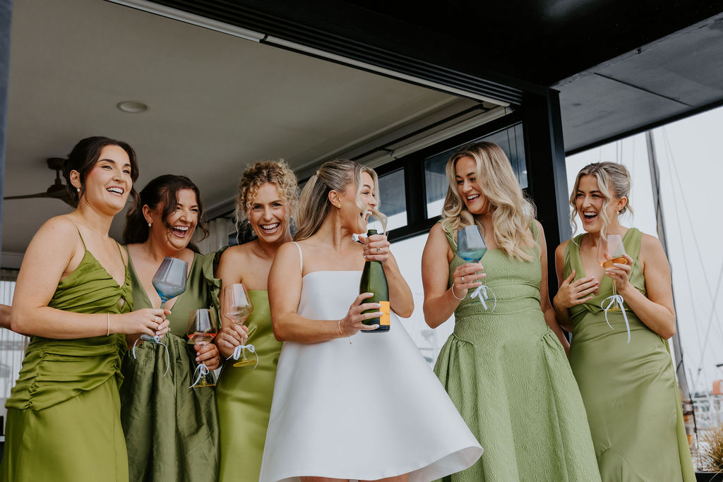 Bride in a white dress surrounded by six bridesmaids in green, toasting with champagne on a balcony.